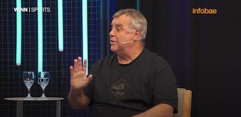 Hombre con cabello gris, camiseta negra con logo, micrófono de solapa y silla de stock marrón, gesticulando. Fondo oscuro con luces azules verticales y copas