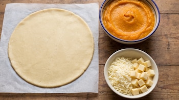 Una masa circular sobre papel de hornear, un tazón con puré anaranjado y un tazón con queso en cubos y rallado sobre una mesa de madera.