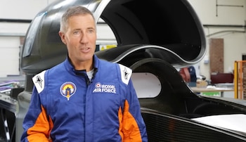 Hombre de mediana edad con traje de piloto azul y naranja con logos de Royal Air Force, de pie frente a un vehículo de competición en un taller