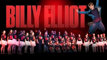 Un joven salta sobre un escenario con el elenco del musical Billy Elliot, incluyendo niños en tutús y adultos, bajo el título rojo "BILLY ELLIOT"
