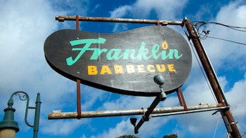 Un letrero ovalado de madera oscura con las palabras 'Franklin' en verde y 'BARBECUE' en amarillo cuelga de una estructura metálica bajo un cielo azul nublado