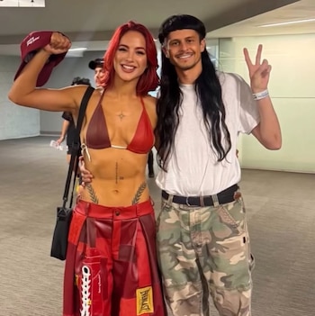 Flor Vigna, de cabello rojo y atuendo rojo, sonríe junto a Oliver Tree, de cabello oscuro, camiseta blanca y pantalones de camuflaje, haciendo el signo de paz