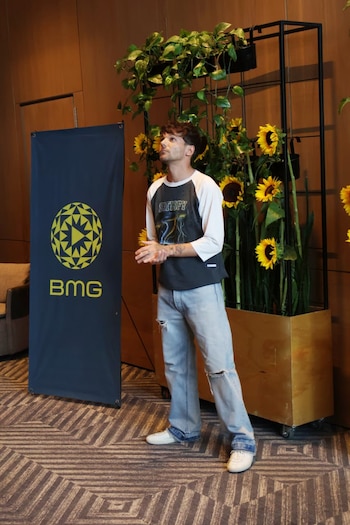 Louis Tomlinson de pie, mirando hacia arriba, con camiseta de béisbol y jeans rotos, frente a un banner de BMG y una estructura con girasoles y plantas artificiales