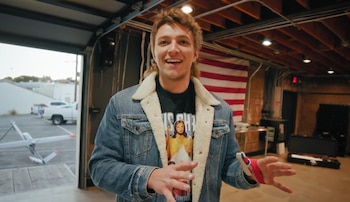 Augustus Doricko sonríe y gesticula en un hangar con una chaqueta vaquera, una camiseta de Jesús y un dispositivo en la muñeca. Detrás, un dron y una bandera de EE. UU