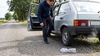 Un hombre junto a un automóvil gris con la puerta abierta mira la rueda trasera, cerca de una botella de plástico aplastada en el asfalto.