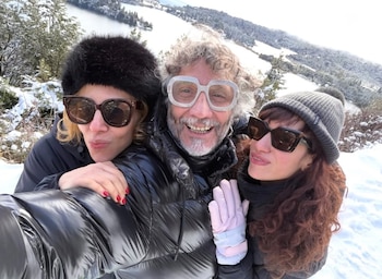 Selfie de tres personas con ropa de invierno, gafas de sol y sombreros en un paisaje nevado, con árboles y un lago al fondo bajo un cielo claro