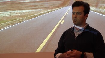 Un hombre de cabello oscuro con suéter azul y camisa a cuadros sostiene un objeto oscuro mientras habla frente a una pantalla con imagen de carretera y campos secos