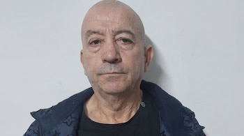 Primer plano de un hombre calvo de mediana edad con barba incipiente, camiseta negra y chaqueta azul oscuro, frente a una pared blanca.