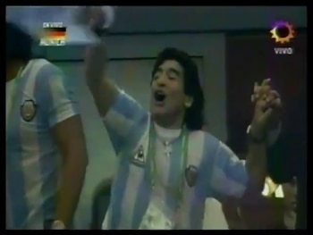 Diego Maradona, con camiseta de Argentina celeste y blanca, con boca abierta y brazos alzados en celebración. A su lado, otra persona. Fondo oscuro. Logotipos de TV visibles