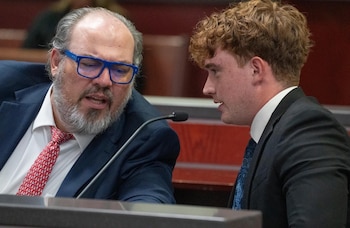 Un hombre con gafas azules de marco grueso y barba habla de cerca con un joven vestido con traje y corbata en la sala de un tribunal