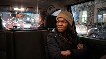 Mujer afrodescendiente sentada en la parte trasera de un taxi, con gorro beige y abrigo oscuro, brazos cruzados, y expresión de enojo e incomodidad. Se ve la lluvia en la ventana y el reflejo del taxista.