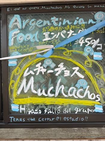 Un cartel vibrante en una ventana con texto "Argentinian Food" y "Muchachos" en blanco y azul, decorado con cintas y banderas argentinas, promocionando comida
