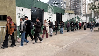 Larga fila de personas camina por una acera de cemento junto a un muro pintado. Al fondo, edificios altos. El cielo está nublado