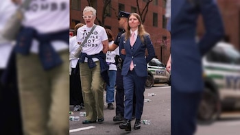 Dos mujeres son escoltadas por un oficial de policía en una calle urbana. Una viste camiseta blanca de protesta, la otra un traje azul y corbata rosa fuerte