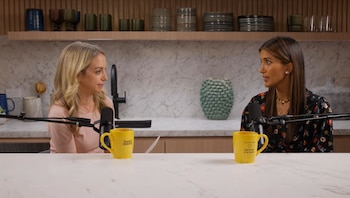 Una mujer rubia y una mujer morena sentadas frente a frente en una encimera blanca con dos micrófonos y dos tazas amarillas. Fondo de cocina moderna