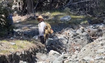 Un hombre con sombrero de paja y mochila camina por un terreno rocoso rodeado de árboles
