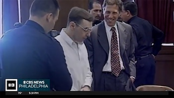Un hombre de cabello corto y gafas en camisa blanca, flanqueado por hombres de traje oscuro, incluyendo uno sonriente con corbata roja y blanca, en una sala de tribunal