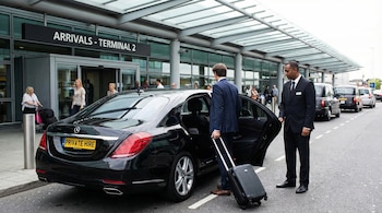Hombre con maleta sube a un coche negro de transporte privado, asistido por un chófer. Se ve la entrada de "Arrivals - Terminal 2" de un aeropuerto.
