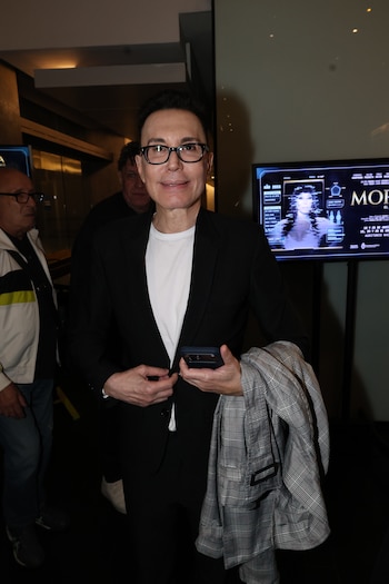 Marcelo Polino, con gafas y blazer negro, sonríe mientras sostiene un teléfono y una chaqueta a cuadros, con una pantalla de Moria Casán al fondo