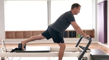 Hombre de mediana edad con camiseta gris y pantalones cortos negros realiza el ejercicio Scooter en un reformer de Pilates, con una pierna extendida hacia atrás.