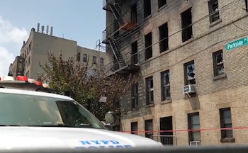Fachada de un edificio de ladrillos con ventanas rotas y áreas ennegrecidas; una escalera de incendios y un coche de policía con luces rojas en primer plano