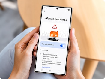Manos de una persona sostienen un teléfono móvil con la pantalla encendida que muestra una aplicación de alertas de sismos y un icono de advertencia