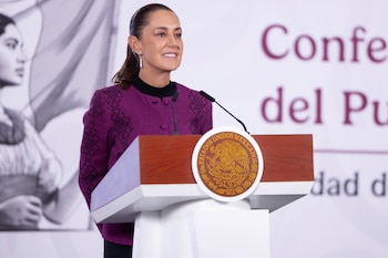 Claudia Sheinbaum Pardo, con una chaqueta magenta y negra, sonríe ligeramente mientras habla desde un podio de madera que muestra el escudo de México