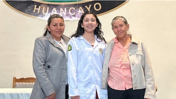 Tres mujeres sonríen frente a una pared clara con un letrero que dice "HUANCAYO". La mujer central lleva un guardapolvo blanco; las otras dos, abrigos