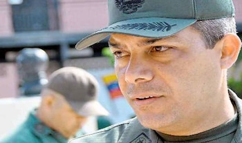 Un hombre con uniforme militar verde y gorra observa hacia un costado mientras otra persona en uniforme permanece al fondo