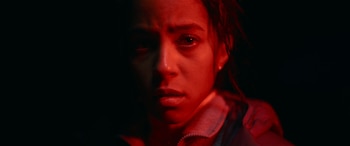 Primer plano del rostro de una joven, con la piel bronceada y el pelo oscuro, iluminado por una luz roja que crea sombras dramáticas en un fondo negro