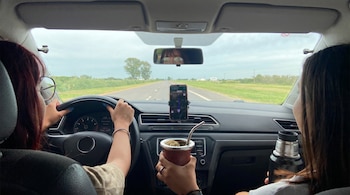 Interior de un automóvil con dos personas en los asientos delanteros. La conductora maneja, la acompañante sostiene un mate y un termo. Un smartphone muestra un mapa GPS en el tablero.