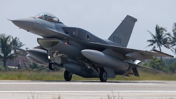 Un avión de combate gris con la matrícula 6728 en la cola despega sobre una pista con palmeras al fondo