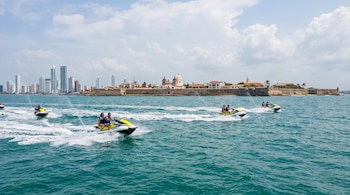 Cinco jetskis con personas navegan en el mar frente a una ciudad con edificios modernos y una antigua muralla colonial bajo un cielo nublado.