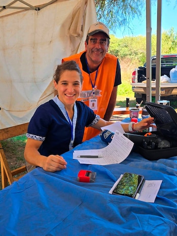 Paula Pareto, vestida de azul y sonriente, se sienta detrás de una mesa con una lona azul, papeles y un teléfono. Un hombre en chaleco naranja la acompaña