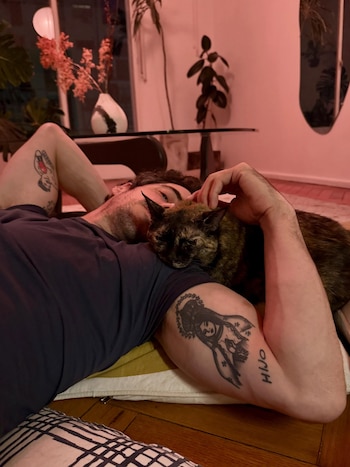 Hombre recostado en el suelo sobre un cojín. Gato atigrado sobre su hombro. Brazos tatuados con figuras y texto. Plantas y luz rojiza al fondo