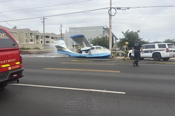 Avioneta blanca y azul con la cola dañada en medio de una calle urbana, con bomberos rociando espuma y un coche de policía cerca