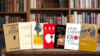 Siete portadas de libros de Anne Carson dispuestos en fila sobre una mesa de madera ante una biblioteca de fondo