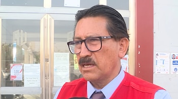 Primer plano de un hombre con anteojos, chaleco rojo, camisa clara y corbata, ubicado frente a una puerta de vidrio con avisos.