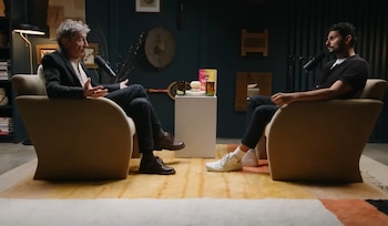 Gabor Maté y un hombre sentados en sillones. Un pedestal con un libro, dos vasos y un objeto decorativo entre ellos. Dos micrófonos, una alfombra