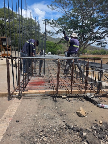 Varios obreros ensamblan una estructura rectangular de varillas de acero sobre el suelo cerca de una carretera con vegetación al fondo