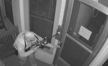 Un oficial de policía uniformado sostiene un arma larga y alcanza el picaporte de una puerta exterior en una toma de cámara de seguridad