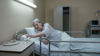 Una mujer mayor, acostada en una cama de hospital con barandales de metal, estira su brazo hacia un vaso de agua y un botón de llamada en la mesa de noche.