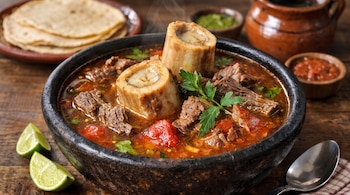 Plato de barro negro con caldo de res, huesos de tuétano, trozos de carne y cilantro, acompañado por rodajas de limón y tortillas.