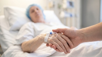 Dos manos se entrelazan; una tiene un catéter intravenoso y la otra la sostiene con cuidado, mientras una persona yace en una cama de hospital al fondo.