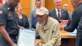 Un hombre mayor de tez oscura con barba blanca, gorra clara y suéter crema recibe un certificado enmarcado de un oficial de policía. Varias personas observan