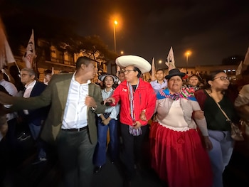 Grupo de personas camina en una calle oscura de noche. Hombre con sombrero blanco y chaqueta roja al centro, mujer con sombrero oscuro y falda roja a su lado, y otros