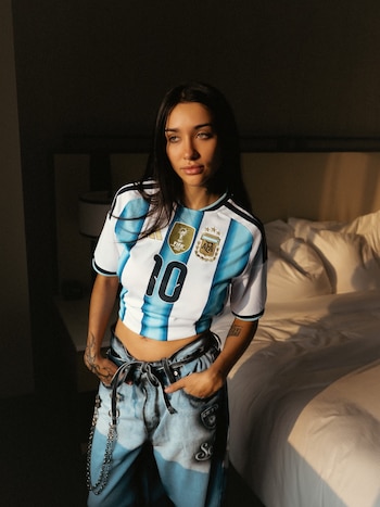 Mujer de cabello oscuro y ojos claros, con camiseta de fútbol argentina número diez y parche de campeón, pantalones vaqueros y cadenas, de pie junto a una cama