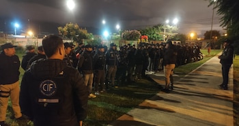 Vista trasera de un oficial con chaqueta del CIF; un numeroso grupo de policías o militares uniformados se alinea en un parque oscuro con farolas encendidas