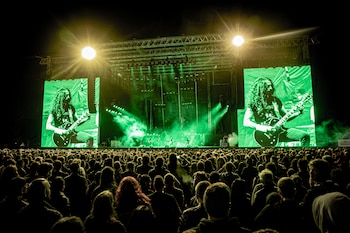 Vista de fundo de uma multidão assistindo a um show de rock em um palco com uma tela gigante mostrando um guitarrista e luzes verdes