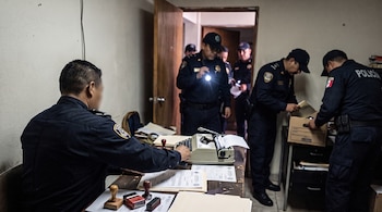 Un hombre con uniforme policial sentado ante un escritorio con máquina de escribir y sellos, junto a policías que revisan la habitación.